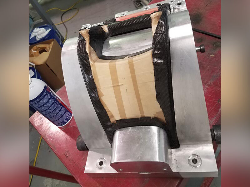 On a red table in a manufacturing space, there is metallic tool being used for layup. Prepreg is on the tool and surrounding a brown plastic coated structure.